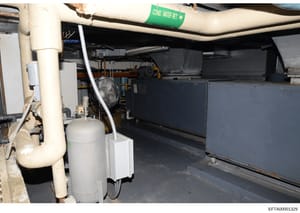Photograph of a basement or mechanical room showing HVAC systems, plumbing infrastructure, and building utilities. A green label marked 'COND. WATER RET.' (condensation water return) is visible on overhead piping. The image documents the interior mec