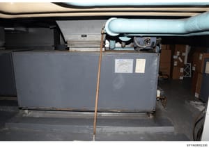 This photograph shows industrial HVAC (heating, ventilation, and air conditioning) equipment installed in what appears to be a basement or mechanical room. The image captures a large gray metal unit with piping, ductwork, and various mechanical compo