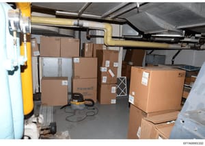 This photograph shows a basement or storage area containing numerous sealed cardboard boxes stacked on shelves and the floor, along with industrial equipment including a yellow vacuum pump, piping, and utility items. The boxes are labeled with shippi