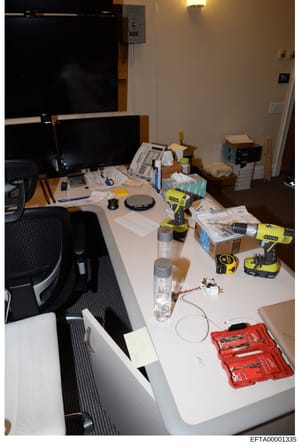 This is an evidence photo from the investigation showing an office or workspace interior. The image captures a desk setup with multiple computer monitors, scattered paperwork, office supplies, and various tools including RYOBI power tools and a red t