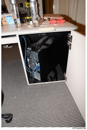 This is a photograph of an office desk area showing computer equipment, power tools, and various items on the desktop and inside an open cabinet. The image appears to be evidence documentation from a search or seizure, with items potentially relevant