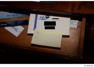 This is an evidence photograph showing a wooden desk surface containing a Blu-ray disc case, what appears to be a television station logo (ABC 22 WNM), and handwritten notations on a yellow sticky note. The image includes redacted text on a white doc