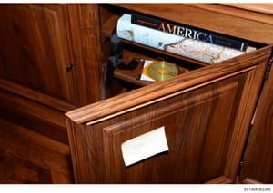This is an evidence photograph showing the interior of a wooden cabinet or desk drawer containing books, maps, and media items. The photo appears to be from a property search or inventory and shows items including an 'America' book by Andres Serrano,