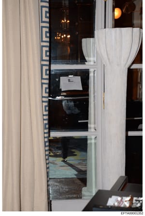 This is an evidence photograph showing the interior of a room taken through a glass door or window. The image captures decorative furnishings including white curtains, patterned upholstery with blue geometric designs, and interior lighting fixtures. 