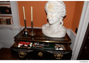 This photograph documents an interior room setting featuring a classical marble bust displayed on an ornate wooden desk with decorative elements. The image shows an elegantly appointed space with orange walls, built-in shelving, and various personal 