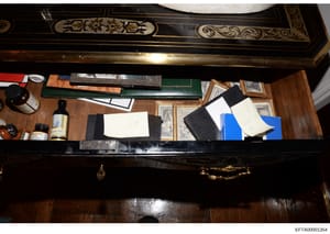 This photograph shows the interior of an ornate desk with decorative inlaid wood and brass details. The desk contains various documents in folders (including green and black leather folders), framed photographs, bottles of spirits, and scattered pape