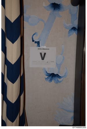 This is an evidence photograph showing an interior wall or surface with decorative blue and white floral wallpaper or fabric pattern. A white evidence marker labeled 'V' is placed on the surface for documentation and scale reference. The image includ
