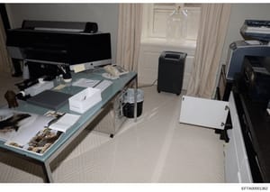 This photograph depicts an office or workspace containing multiple computer workstations, printers, and various personal items and photographs scattered on a glass-topped desk. The image appears to be evidence documentation from a property search or 