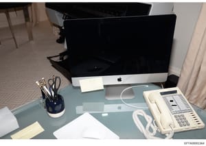 This is an evidence photograph depicting a modern office workspace with contemporary computer equipment. The desk features an Apple iMac desktop computer, wireless mouse, telephone system, and standard office supplies. The image documents the physica