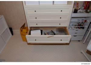 This is an evidence photograph showing an interior bedroom scene with a white wooden dresser with multiple drawers. The bottom drawer is open, revealing storage contents including white items, DVDs or media cases, and other personal effects. A yellow