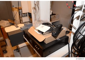 This photograph documents seized office equipment and electronics, including computer peripherals, cables, and a stapler with a handwritten label marked 'Exhibit A'. The image appears to be taken during an evidence collection or search operation, sho