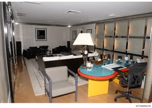 This is a photograph of a contemporary office interior featuring a prominent desk with a turquoise top and yellow base, modern black seating furniture, framed artwork on walls, and professional office equipment. The space appears to be part of a resi