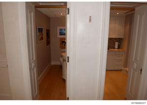 This is a photograph showing interior hallways and adjoining rooms of a residential property, including views of a bathroom with toilet visible, a kitchenette area with cabinets and countertop, and multiple doorways with white frames. The image appea
