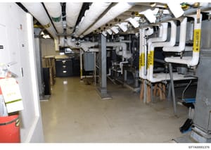 This photograph shows the basement mechanical infrastructure of a property, featuring HVAC systems, plumbing pipes, heating equipment, and various utility installations. The image documents the building's mechanical systems and may be relevant to pro