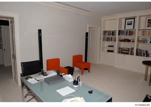 This is a photograph of a spacious, contemporary interior room featuring a glass desk workspace, built-in bookshelves, and modern furniture including orange accent chairs. The room appears to be part of a residence or office property documented durin