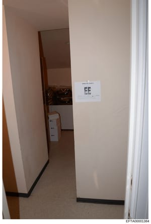 This is a photograph of an interior room or hallway, showing a doorway with white walls, carpeted flooring, and a white label marked 'EE' affixed to the wall. The image appears to be evidence documentation from the investigation, with a Bates number 