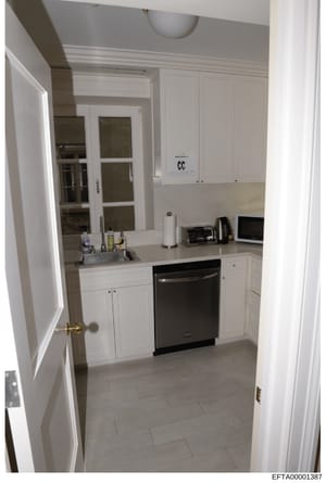 This is a color photograph of a modern, compact kitchen with white cabinetry, stainless steel appliances, and light tile flooring. The image shows a well-maintained residential kitchen space with standard furnishings and countertop items. The photo i