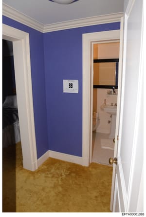 This photograph shows the interior of a residential room with distinctive blue walls, white trim, and tan carpet. The image captures a hallway or corridor area with doorways leading to what appears to be a bedroom (on the left) and a bathroom with vi