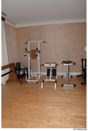 This is an evidence photo showing a residential gym space containing multiple pieces of exercise equipment including stationary bikes, weight machines, and other fitness apparatus. The room features light wood flooring, beige textured walls, and crea