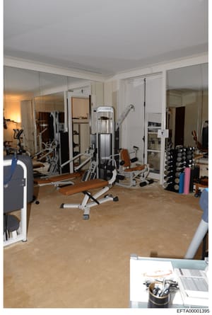 This photograph depicts a residential fitness room equipped with extensive exercise machinery, weight training equipment, and mirrors. The room contains multiple pieces of commercial-grade fitness equipment including what appears to be a stationary b