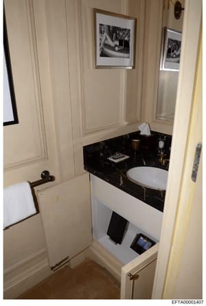 This is a photograph of a luxurious bathroom interior, showing a modern vanity with black marble countertop, white circular sink, framed black-and-white artwork on cream-colored walls, and brass fixtures. The image appears to be evidence documentatio