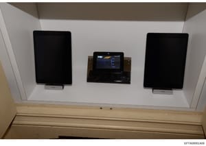 This photograph shows an interior wall-mounted display system consisting of three screens: two larger tablet-style devices on either side (marked 'belkin') and a central control panel with an illuminated interface displaying what appears to be a navi
