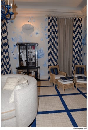 This is a photograph of a luxuriously decorated bedroom featuring a blue and white color scheme with high-end furnishings. The room includes a curved white upholstered bed, custom wallpaper with blue floral motifs, bold patterned curtains, a black di