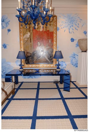 This is a photograph of an elegantly decorated interior room, featuring a sophisticated display with a blue Murano glass chandelier, ornate gold-framed mirror, portrait painting, and symmetrical blue table lamps. The room showcases high-end furnishin