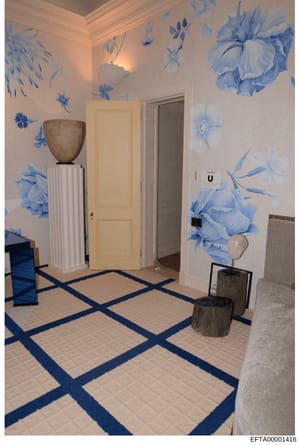 This photograph shows an elegantly decorated hallway interior featuring distinctive blue floral wallpaper, a geometric blue and white tile floor pattern, classical architectural details including crown molding, and decorative elements including ceram