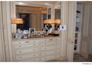 This photograph documents a high-end residential dressing area or master bedroom vanity, featuring custom cabinetry, multiple mirrors, decorative lighting, and personal items including framed photographs and grooming products. The image appears to be