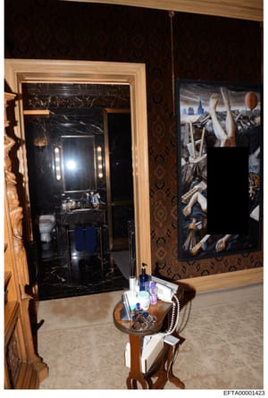 This is a photograph of an opulent bedroom interior, showing a black marble bathroom with illuminated vanity mirror, dark patterned wallpaper, and a framed classical artwork depicting religious or mythological figures. The room contains luxury furnis