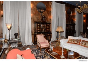 This photograph shows an elegantly decorated interior room featuring high-end furniture, including a pink upholstered chair, white sofa with decorative pillows, dark wood cabinetry with inlaid details, multiple mirrors, brass and crystal lighting fix