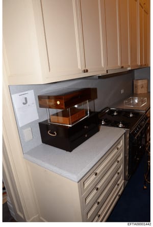 This photograph shows a kitchen counter area with custom cabinetry and appliances. A black box with wooden drawers and brass handles sits on the counter, appearing to be evidence or a seized item marked with a 'J' label. The image documents the inter