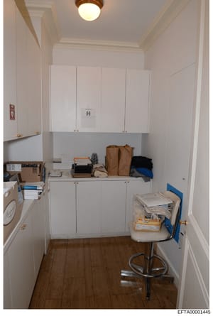 This photograph shows a utility or storage room, possibly from one of Epstein's properties, featuring white cabinetry, wooden flooring, and various items stored on shelves. The room contains cardboard boxes, brown paper bags, clothing, documents, and
