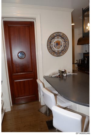 This is an interior photograph of what appears to be a high-end residential kitchen or dining area, showing a dark countertop, white modern seating, a mahogany door with circular window, and decorative heraldic plate on the wall. The photograph is pa