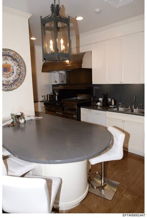 This is a photograph of a modern, upscale kitchen interior featuring a curved gray-topped island with white seating, professional-grade black appliances, white cabinetry, and decorative elements including a hanging lantern and ornamental plate. The p