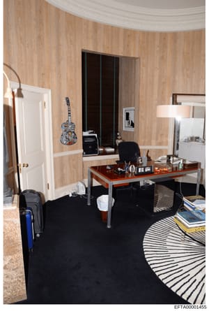 This is a photograph of a furnished bedroom featuring a work desk, decorative elements, and personal items. The room displays wood-paneling walls, a curved ceiling detail, and various furnishings including a desk with office supplies, a black office 