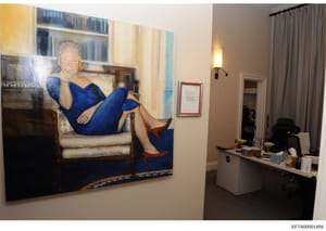 This photograph shows an interior office or residential space containing a large portrait painting of a woman in a blue dress seated on a chair. The space includes modern furnishings, shelving, desk areas, and curtained windows. The image was likely 