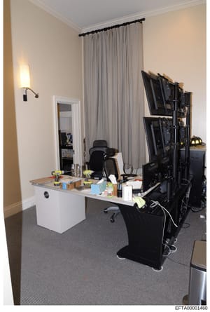 This photograph shows an interior office or workspace room containing computer equipment, desks, and furnishings. The room features a white desk with a mirror, multiple computer monitors on a stand, office chairs, and various personal items. This app