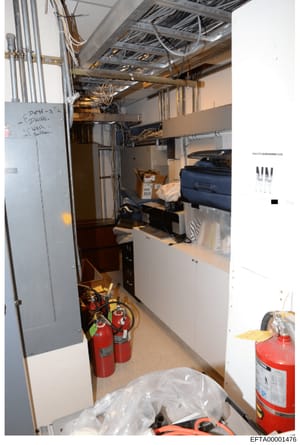 This photograph shows a basement utility room or mechanical space containing HVAC equipment, electrical infrastructure, fire extinguishers, and miscellaneous storage items. The space includes exposed ductwork, piping, electrical conduit, and handwrit