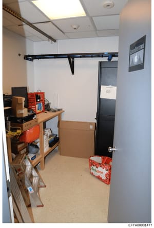 This is a photograph of a small interior room or closet space containing various electronic equipment, storage boxes, and networking infrastructure. The room features wooden shelving units with red storage bins, a black door, and professional-grade o