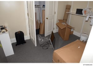 This is an evidence photograph showing an interior room containing multiple cardboard boxes, a black suitcase, rolled materials, and various storage items. The room appears to be a closet, storage area, or hallway with white doors, beige walls, and g