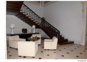 This photograph depicts the interior of a luxury residence, showing an elegant foyer with a dark wood grand staircase featuring ornate wrought iron railings, cream-colored upholstered seating furniture, decorative sculptures and art objects, and orna