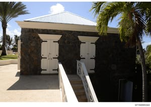 This is a color photograph of a small auxiliary building or cabana structure featuring dark stone columns, cream-colored doors, and a peaked metal roof. The building appears to be located on a tropical estate with palm trees visible in the surroundin