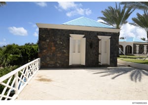 This photograph shows a modern residential outbuilding or accessory structure featuring dark stone walls, white-trimmed doors, and a turquoise metal roof. The building is part of a larger property complex visible in the background, set in a tropical 