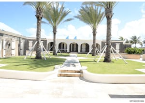 This is a photograph of the exterior courtyard and main entrance of a large residential property in a tropical location. The image shows a modern architectural design featuring multiple tall palm trees, white concrete pathways, arched architectural e