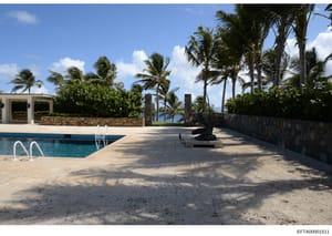 This photograph shows the exterior grounds and swimming pool area of a tropical oceanfront property, featuring a rectangular in-ground pool, pool house structure, manicured landscaping with palm trees, and ocean views. The image appears to be part of