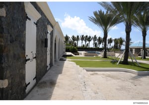 This photograph shows an outdoor area of a luxury property featuring a swimming pool, manicured grounds with palm trees, and a row of guest houses or pool cabanas with white doors and dark stone/slate exterior walls. The landscaping and architectural