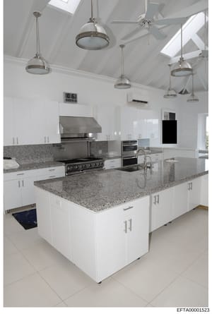 This is a photograph of a contemporary kitchen space featuring white cabinetry, granite countertops, stainless steel appliances, and professional-grade lighting. The image shows a large central island with seating area and multiple pendant lights sus