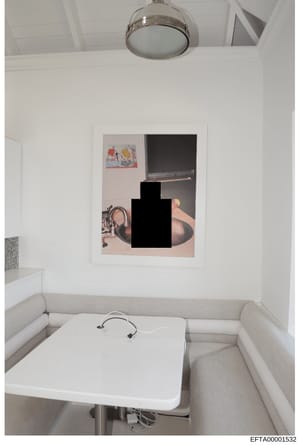 This is a photograph of a small, minimalist room with white walls and ceiling-mounted lighting. The room contains a white table or platform with headphones, beige seating along the walls, and a framed artwork on the wall with a large redacted black s
